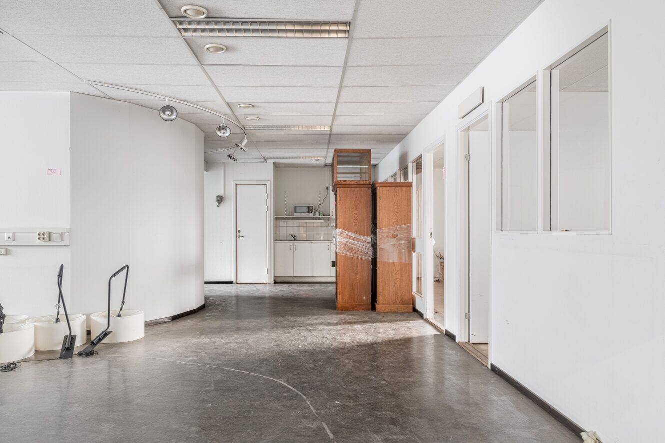Tomt kontorsutrymme med mörkt golv, vita väggar, städredskap till vänster och plastinpackade träskåp nära ett litet pentry längst bak.