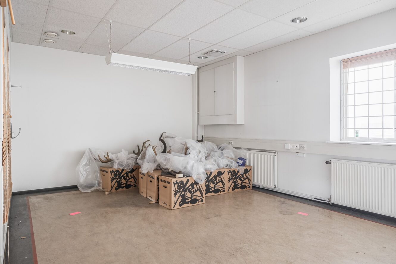 Ett tomt rum med kaklat tak och bart golv, innehållande flera kartonger och horn täckta av plast nära ett fönster med galler.