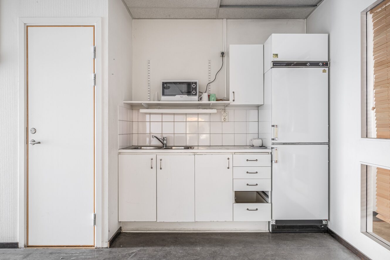 Ett litet pentry med diskho, mikrovågsugn, kylskåp, skåp och delvis öppna lådor; vitt kakel och vita väggar, med en stängd dörr och fönster på sidorna.