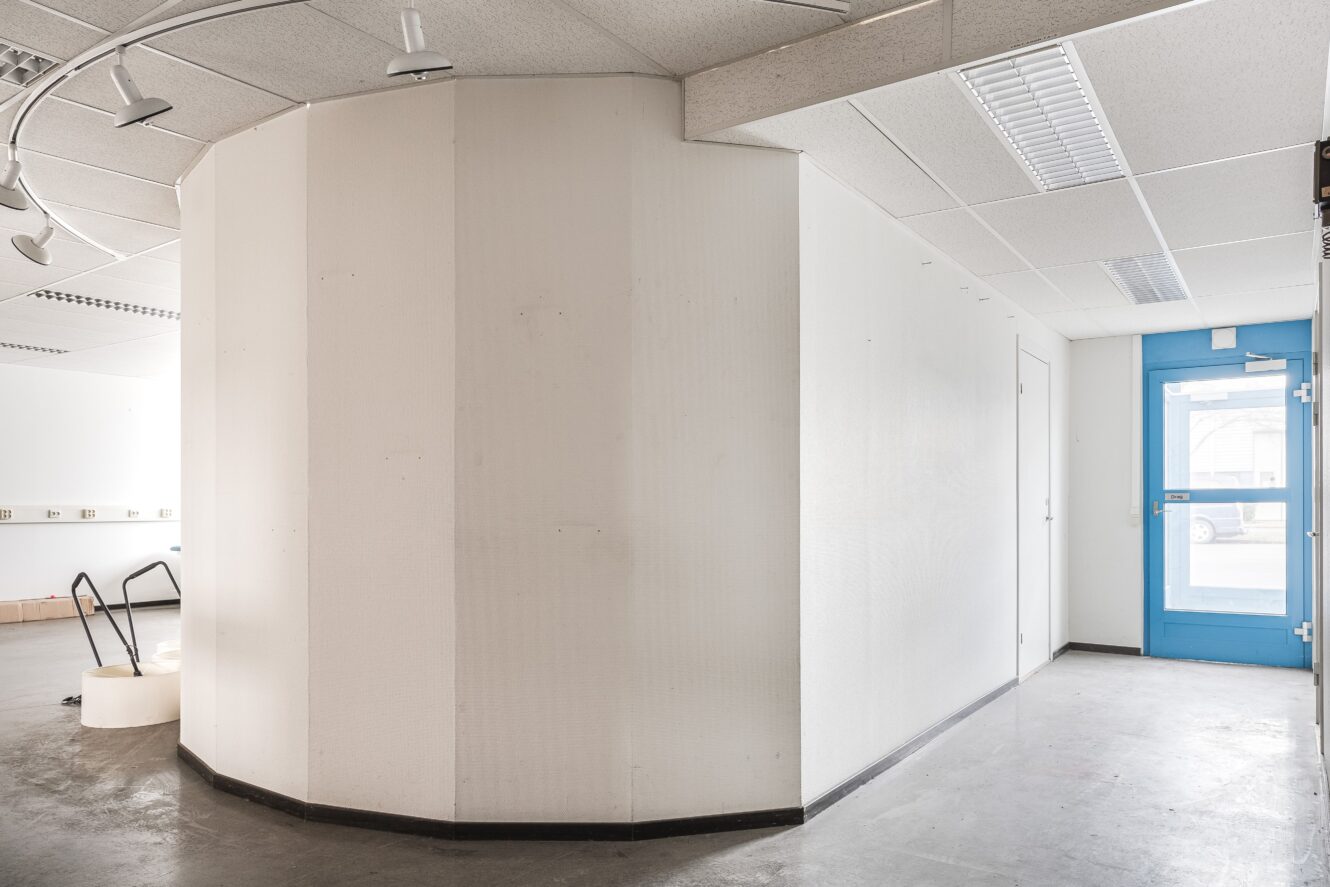 En vit, vinkelformad innervägg med spårbelysning, grått golv och en blå dörr i en i stort sett tom kommersiell lokal.