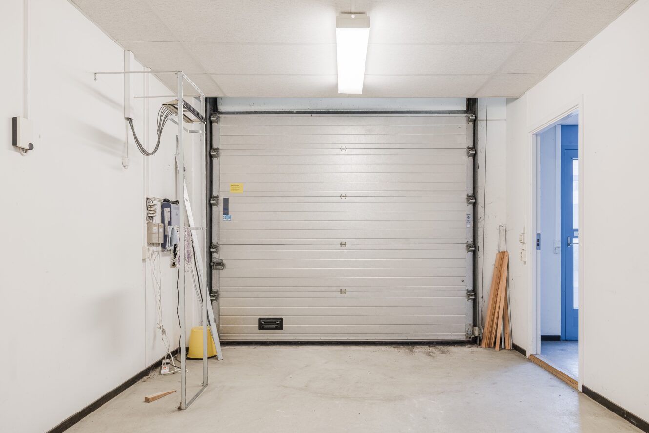 Ett tomt garage med vita väggar, en stängd rullport, ett lysrör i taket, frilagda ledningar och en dörröppning på höger sida.