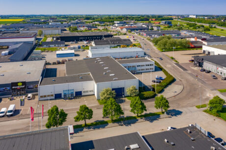 Flygfoto över ett industriområde med lagerlokaler, kontorsbyggnader, parkerade fordon och träd längs gatan en klar dag.