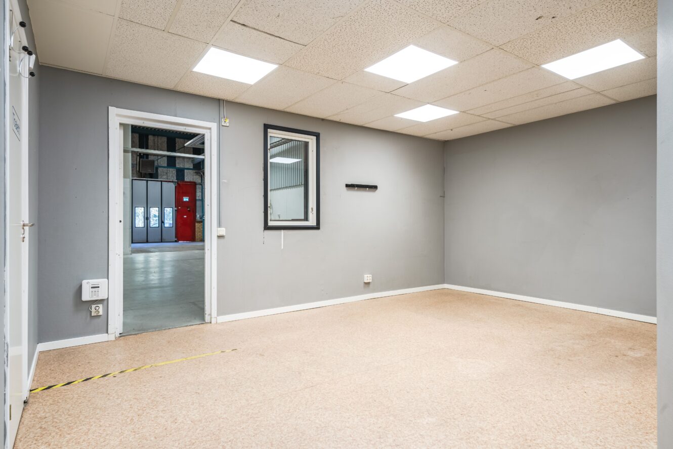 Ett tomt rum med grå väggar och beige golv, med ett fönster mot ett annat rum och en dörr som leder ut till ett större utrymme där man i bakgrunden kan se varuautomater.