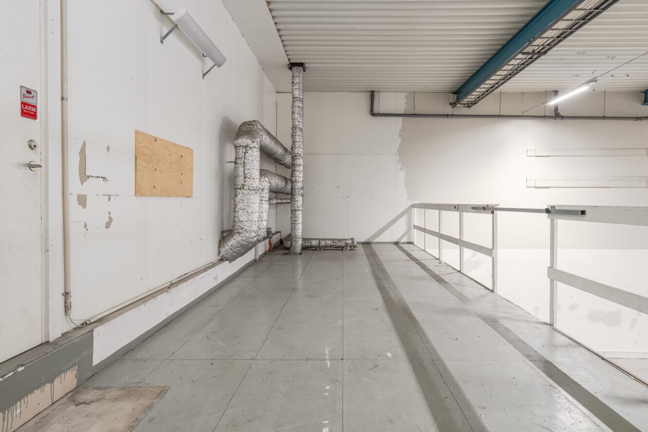 Industriell mellanvåning med grått golv, synliga ventilationsrör insvepta i isolering, vita väggar, räcken av metall och takmonterade lysrör.