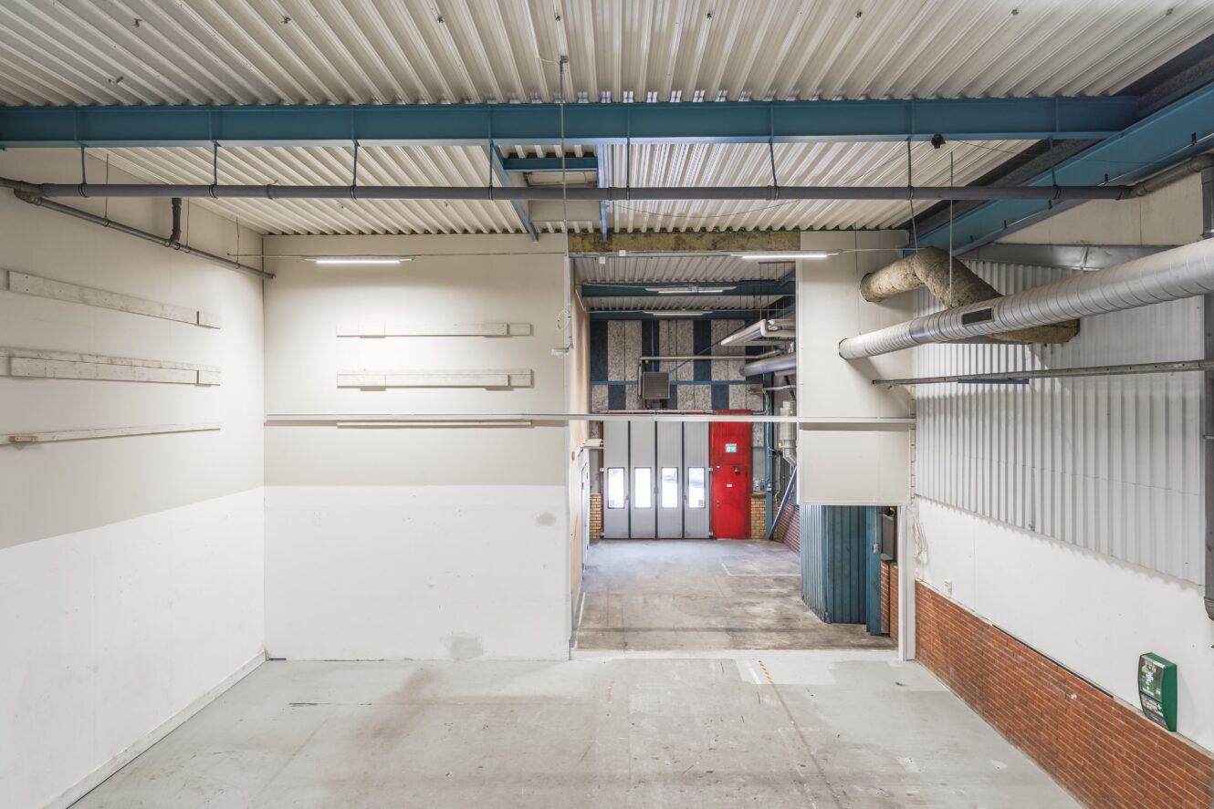 Interiör i ett tomt industrilager med synliga rör, högt tak, betonggolv och en stor garageport längst bort.