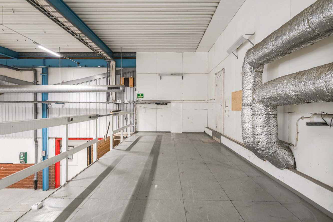 En industriell mellanvåning med metallräcken, vita väggar, isolerade rör och ett grått golv. En skylt med texten ”Nödutgång” och den synliga takkonstruktionen syns tydligt.