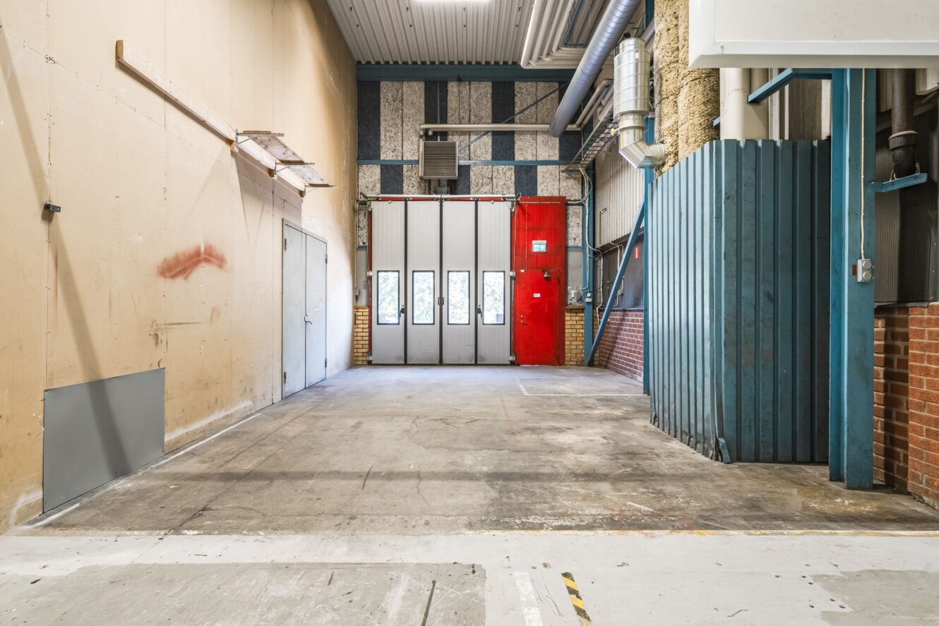 Interiör i ett industriellt garage med betonggolv, stor takskjutport med fönster, röd metalldörr, synliga rör och väggar byggda av olika material.