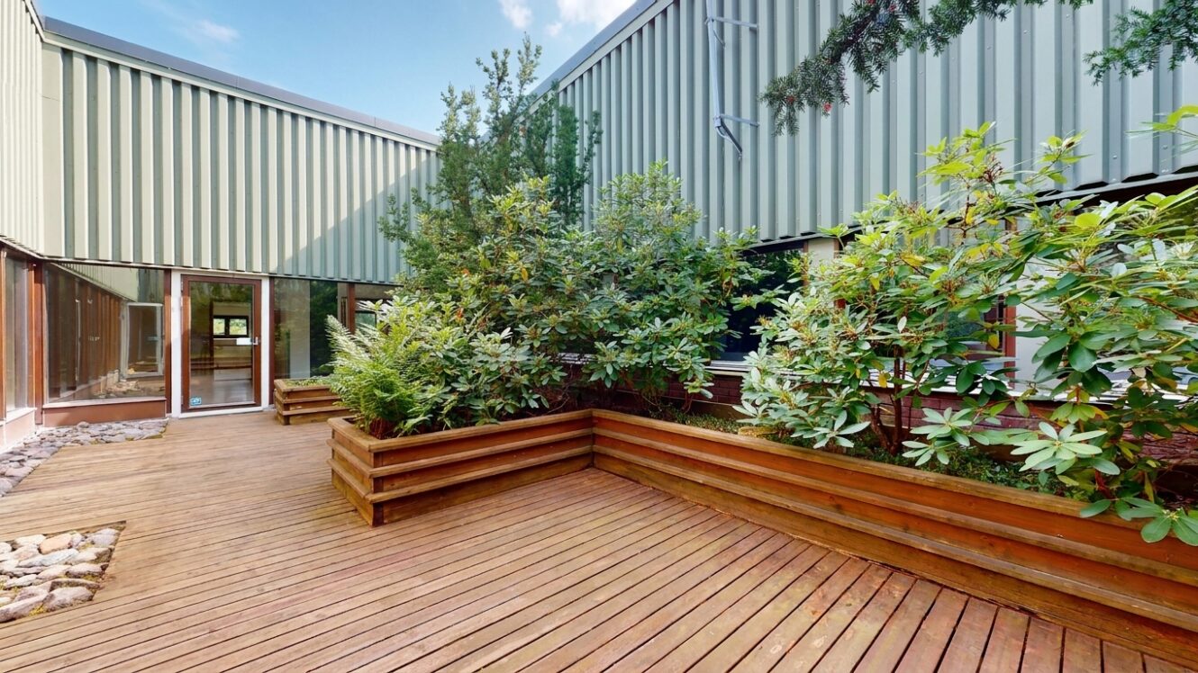 En innergård med träterrass, där det finns en stor upphöjd planteringslåda med buskar och en intilliggande byggnadsingång.