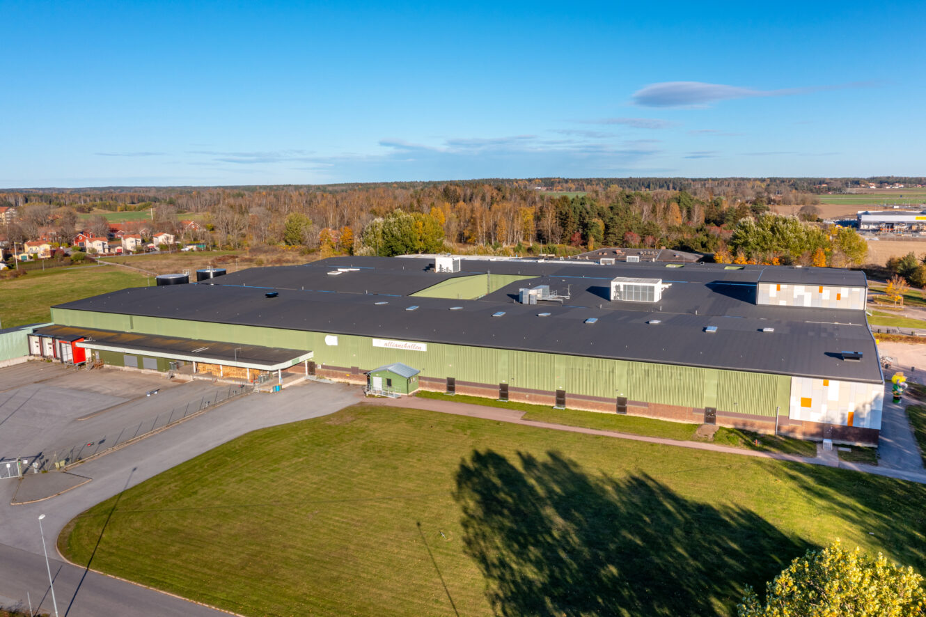 Flygfoto över en stor, rektangulär lagerbyggnad omgiven av gräs, träd och landsbygd under en klarblå himmel.