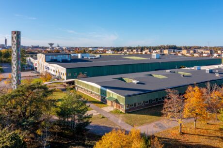 Ett stort grönt industriellt lagerkomplex omgivet av höstträd, med en hög, dekorerad skorsten och en klarblå himmel ovanför.