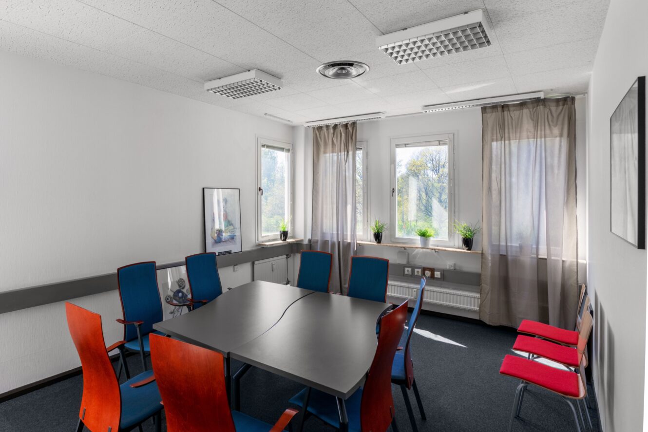 Ett litet mötesrum med ett svart bord, åtta stolar, stora fönster med skira gardiner och flera krukväxter på fönsterbrädorna.