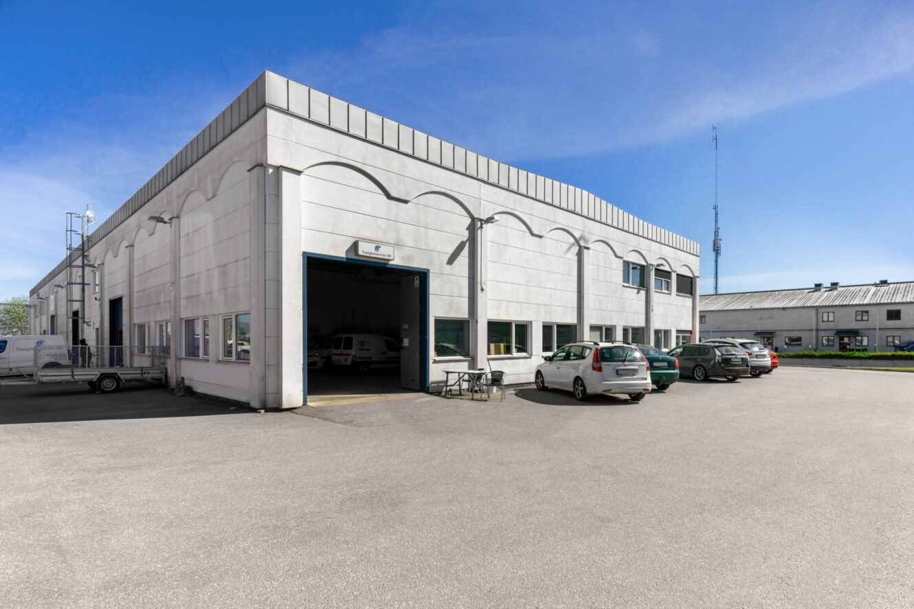 En vit industribyggnad med flera parkerade bilar framför, med en stor garageport och flera fönster en solig dag.