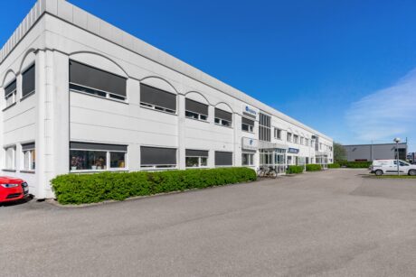 En modern vit kontorsbyggnad i två våningar med stora fönster, gröna buskar längs framsidan och en klarblå himmel ovanför. En röd bil och en vit skåpbil är parkerade i närheten.