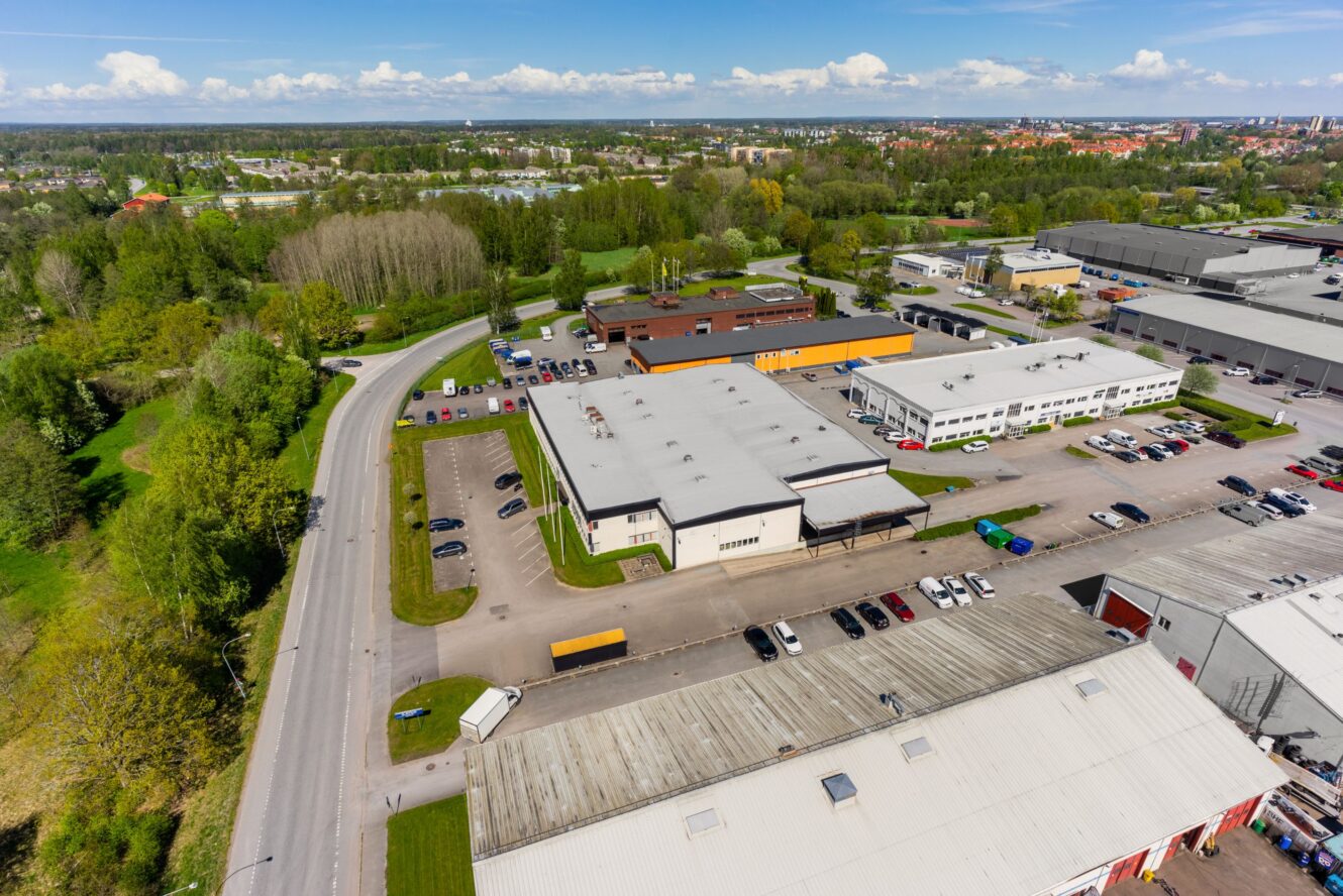 Flygbild över ett industriområde med lagerbyggnader, parkerade bilar, omgivande träd och en väg som slingrar sig genom landskapet.