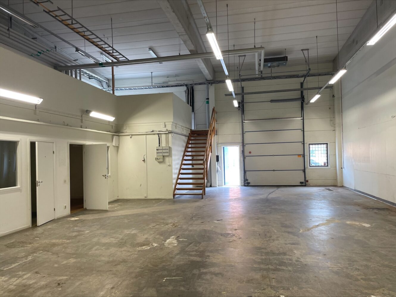Ett tomt industrilager med betonggolv, vita väggar, takbelysning, en trappa till ett litet loft och stora garageportar.