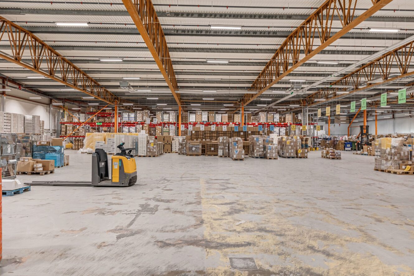 Stor lagerlokal med hyllor, staplade lådor och pallar; en gul elektrisk pallbock står parkerad i förgrunden på ett betonggolv.