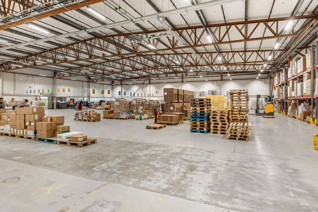 Stor lagerlokal med staplade kartonger och pallar, hyllor längs väggarna och industriell belysning ovanför.