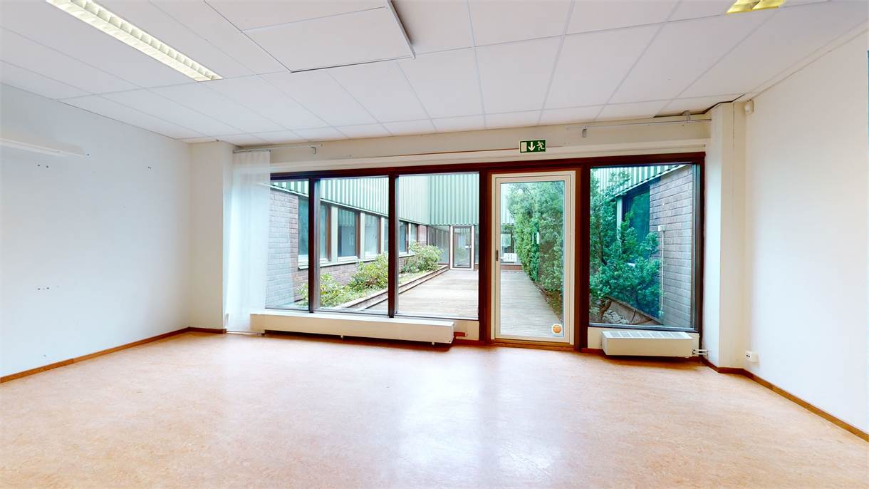 Tomt rum med vita väggar, ljusbrunt golv, stora fönster och en glasdörr med utsikt över en smal innergård med grönska och en gångväg.