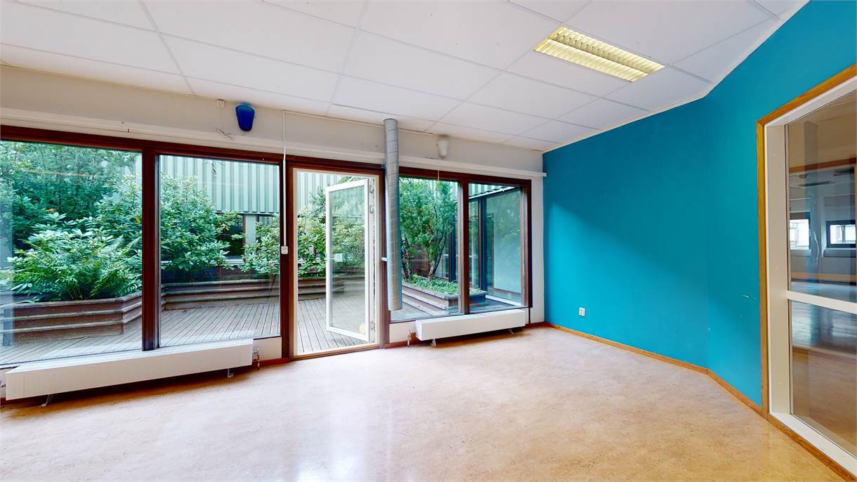 Tomt rum med en blå accentvägg, stora fönster, en öppen dörr som leder till en uteplats med grönska och ett beige golv.