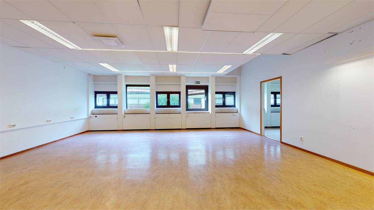 Tomt rum med vita väggar, beige golv, taklampor, tre fönster och en öppen dörröppning till höger.