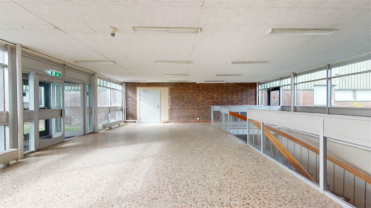 Tom korridor med terrazzogolv, accentvägg i tegel, lysrör i taket, trappräcke till höger och stora fönster till vänster.