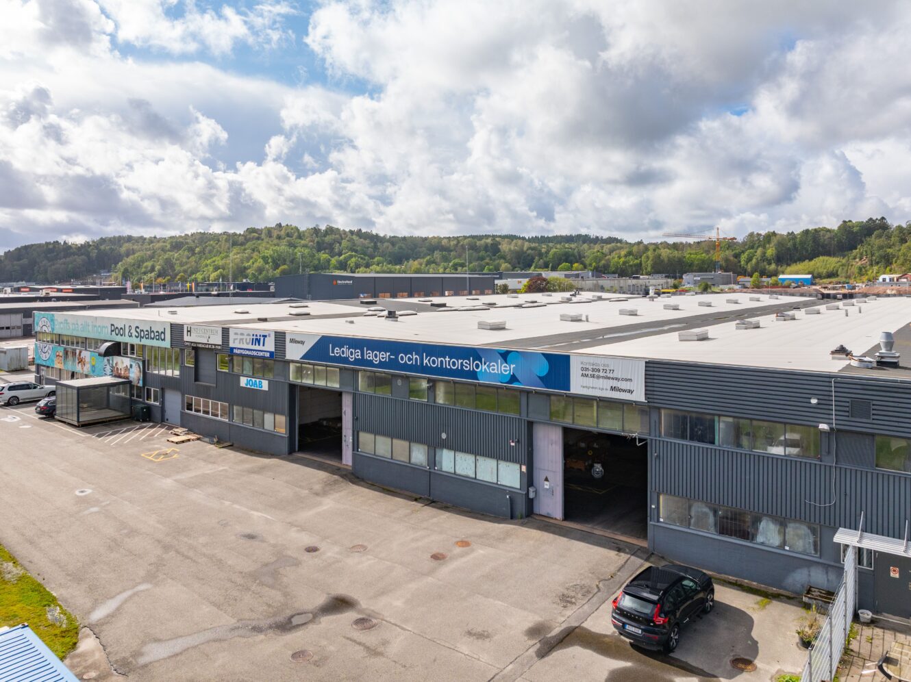 En stor lagerbyggnad med flera ingångar och parkeringsplatser, med en skylt som visar lediga kontors- och lagerutrymmen. Träd och kullar syns i bakgrunden.