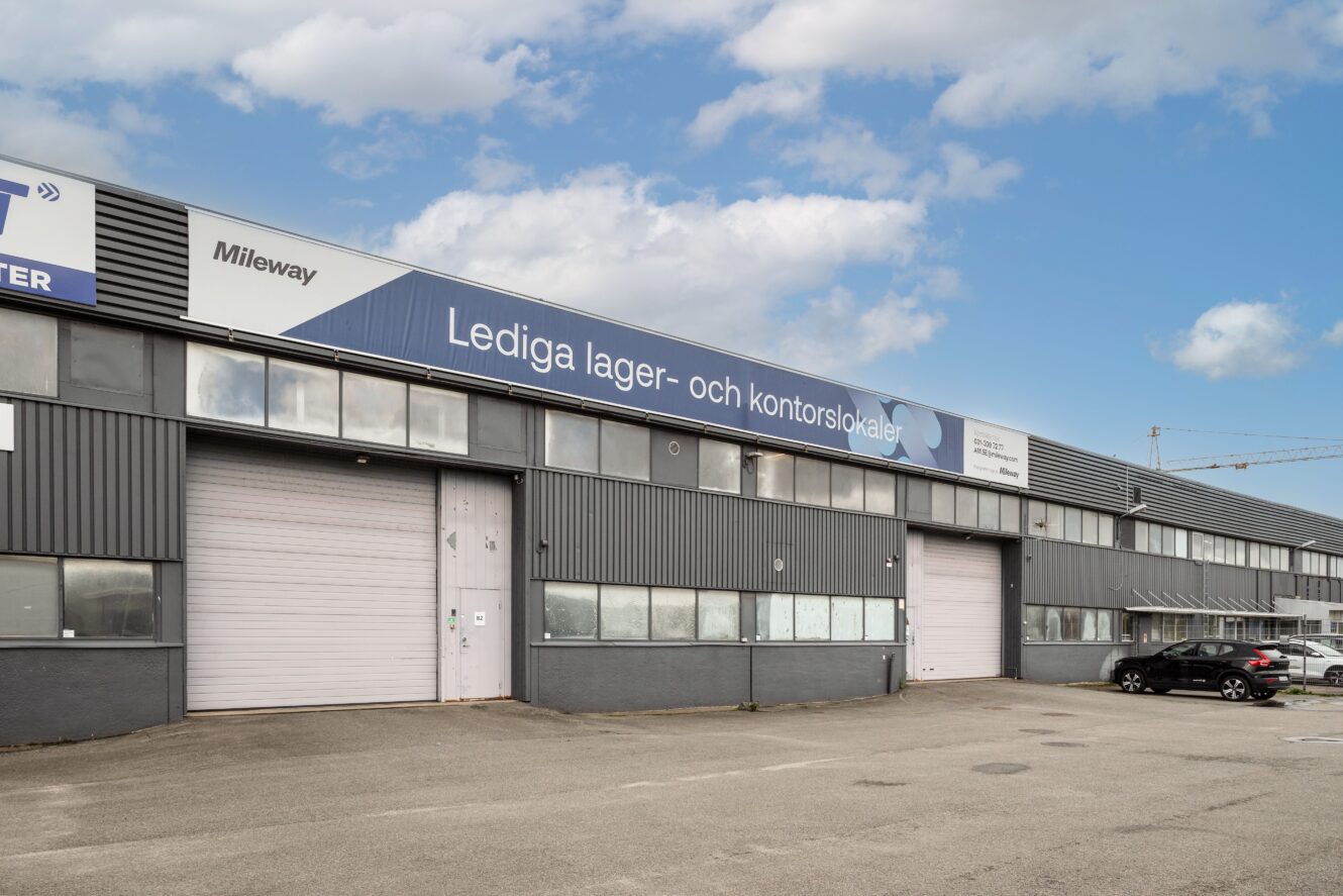 En grå industribyggnad med stora garageportar och en skylt på svenska som annonserar lediga lager- och kontorslokaler; flera bilar står parkerade utanför.