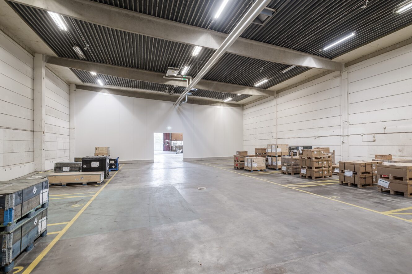 Interiör från en rymlig lagerlokal med högt i tak, vita väggar, takbelysning och staplar av pallar och lådor längs ena sidan. En öppen dörröppning syns i bortre änden.