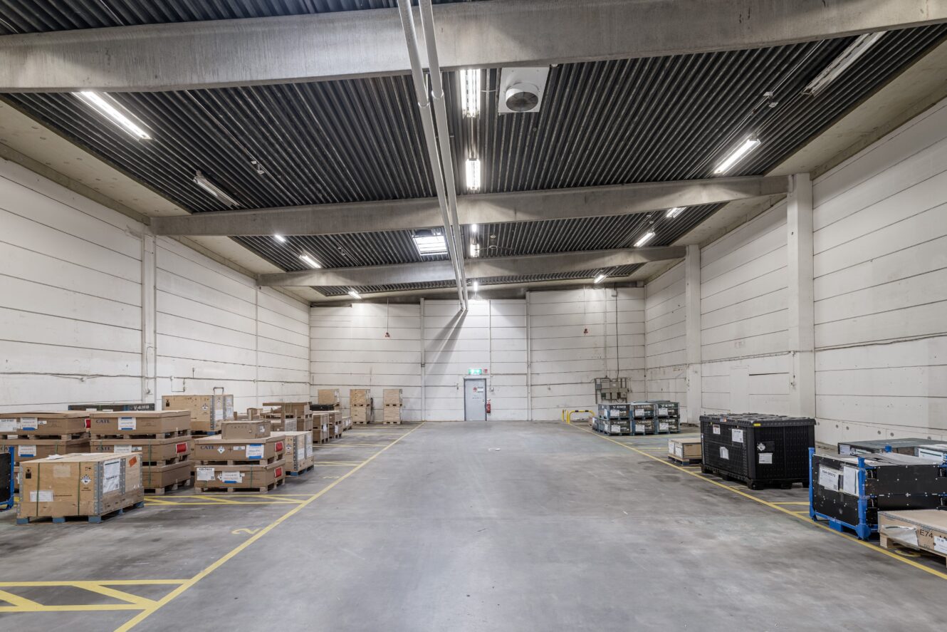 Stor, tom lagerlokal med högt i tak, betonggolv och minimal takbelysning; flera pallar och lådor är placerade längs väggarna.