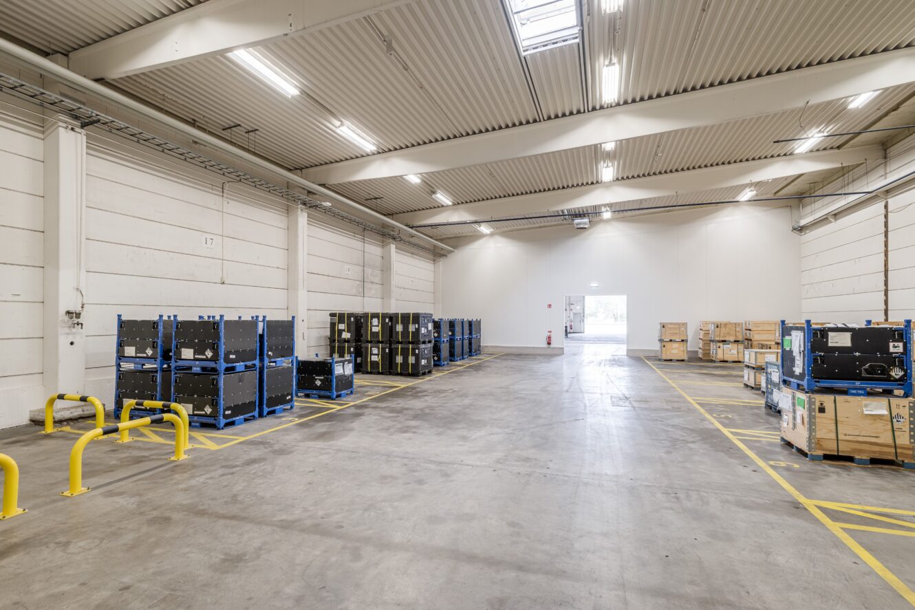 Rymlig lagerlokal med staplade pallar och containrar, högt i tak, stark belysning och tydligt markerade gula säkerhetslinjer på golvet.