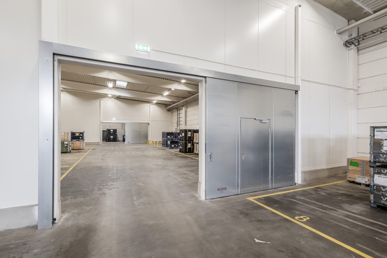 Stor industrilagerlokal med öppen skjutdörr i metall, synliga pallar och material inuti, betonggolv och högt i tak med lysrörsbelysning.