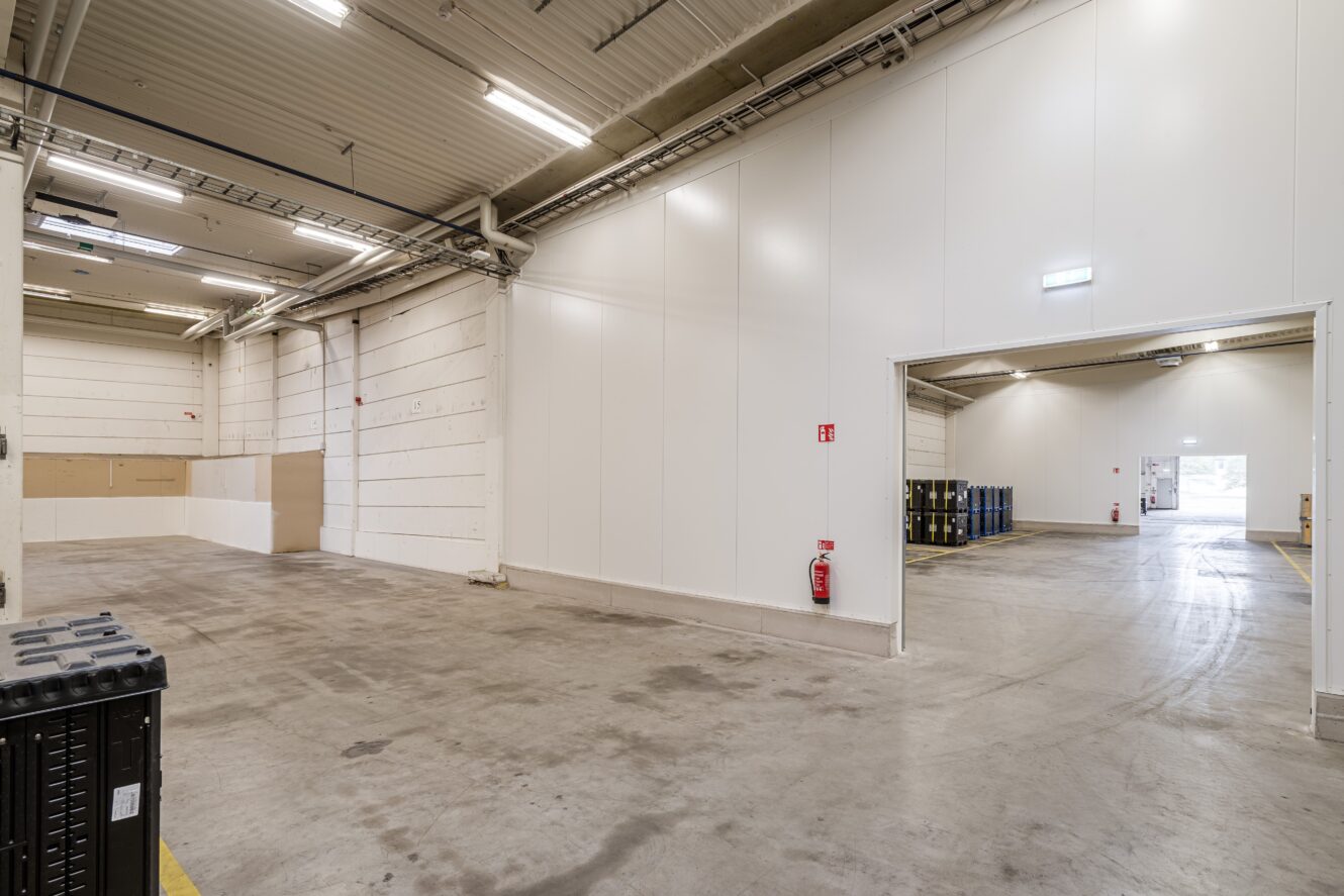 Stor, tom industrilokal med betonggolv, vita väggar, högt i tak, lysrörsbelysning och en synlig brandsläckare monterad på väggen.