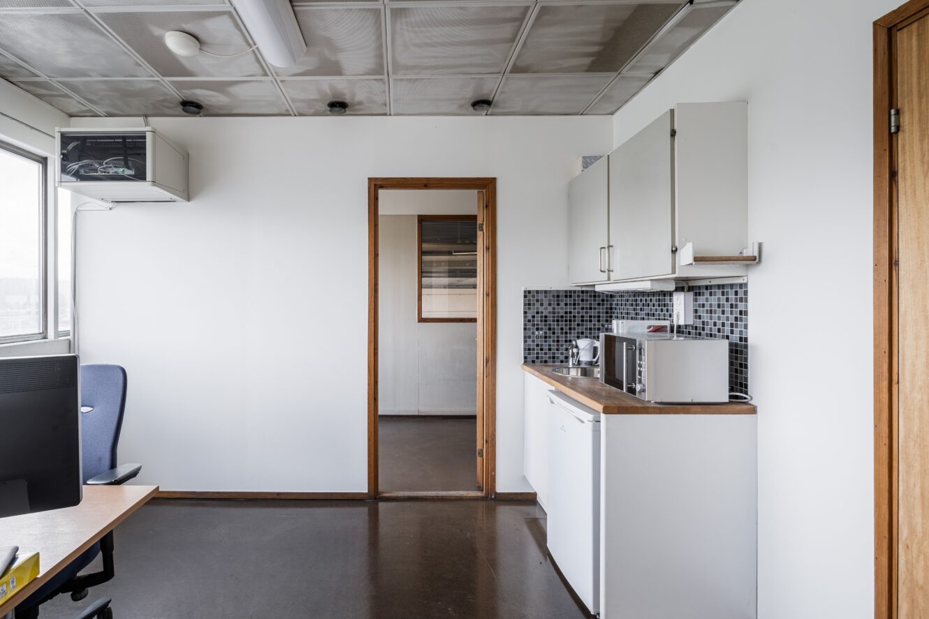 Litet kontorskök med vita skåp, minikylskåp, mikrovågsugn och stänkskydd av mosaikplattor, intill en arbetsplats med stol och skrivbord nära ett fönster.