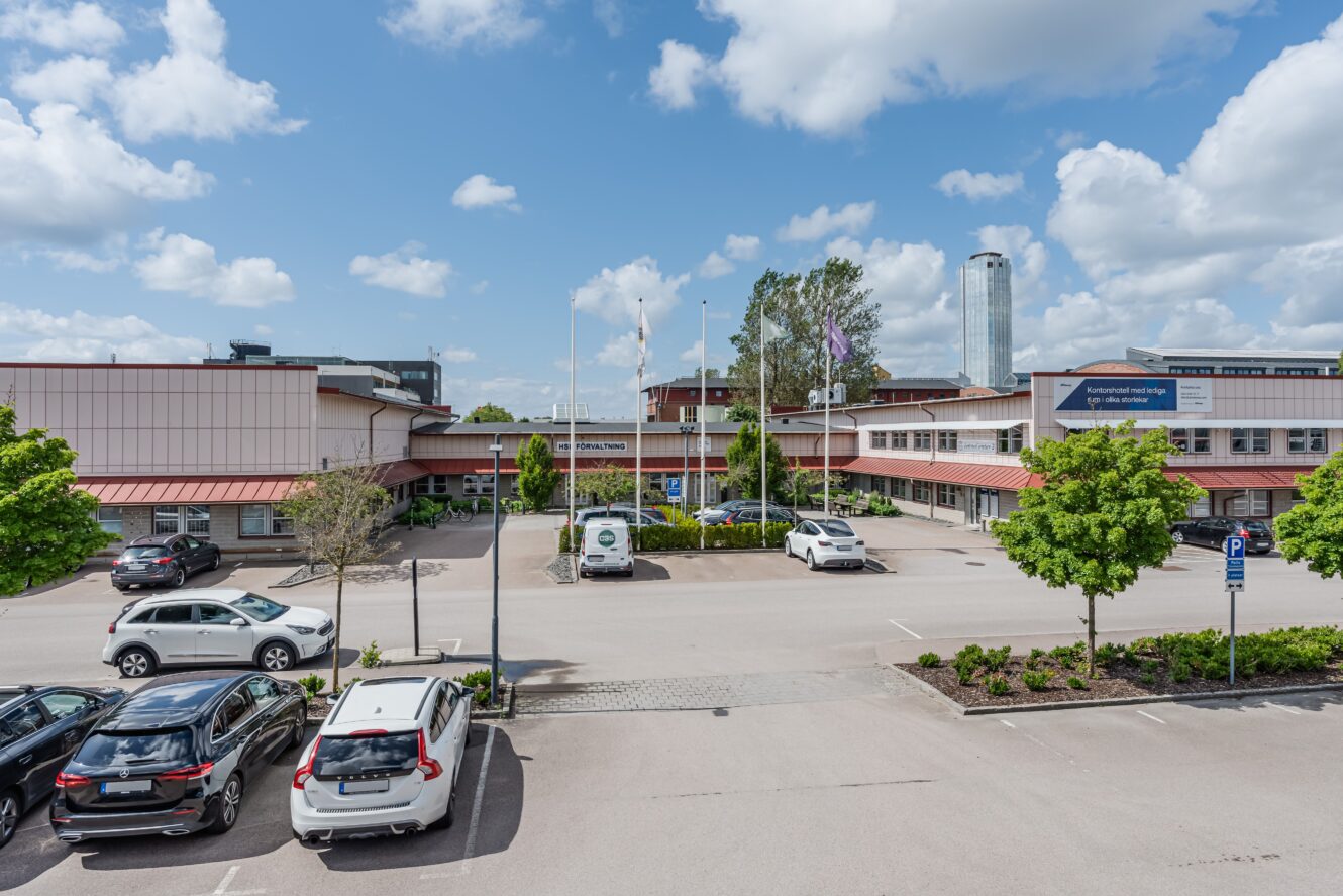 En parkeringsplats med flera bilar ligger framför en låg byggnad med rött tak, flaggstänger, träd och en delvis molnig himmel ovanför.