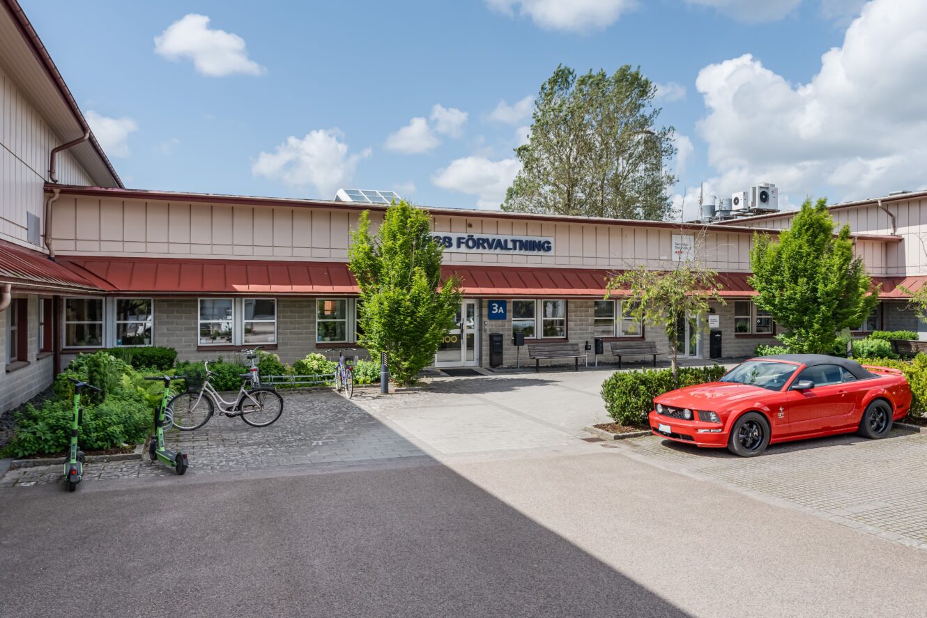 En röd sportbil, en cykel och två elsparkcyklar står parkerade utanför en beige byggnad med beteckningen SB Förvaltning en solig dag.