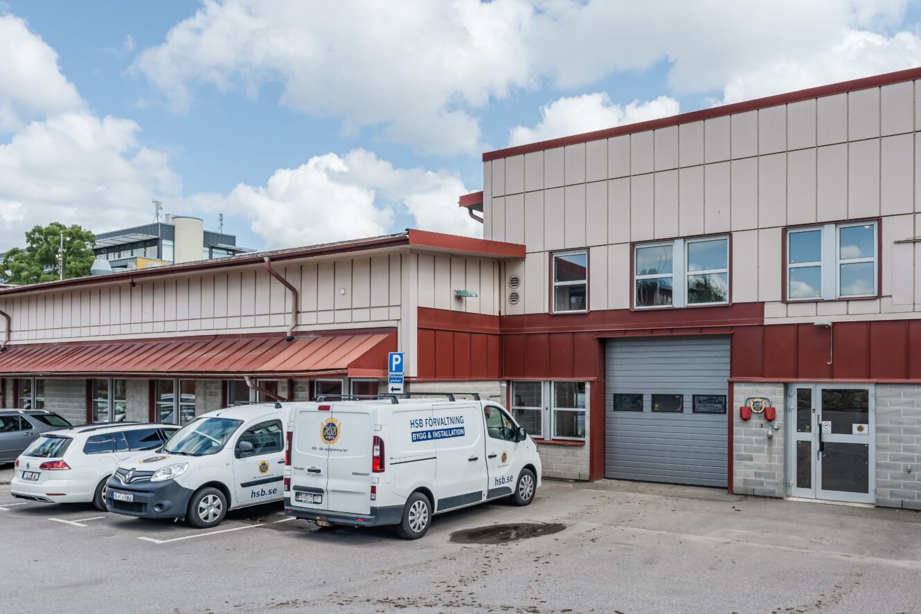 En parkeringsplats med tre fordon, däribland en vit skåpbil märkt HSB Parkering Västra Mälardalen, ligger framför en beige och röd industribyggnad med stora garageportar.