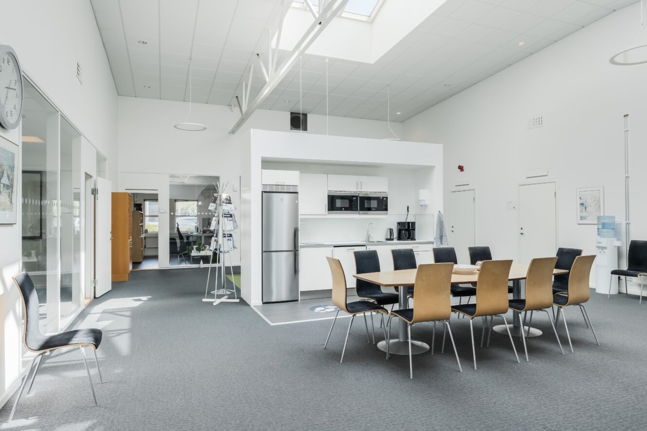 Modernt kontorskök och matplats med bord, stolar, kylskåp, mikrovågsugn och kaffebryggare; ljust utrymme med vita väggar och stora fönster.