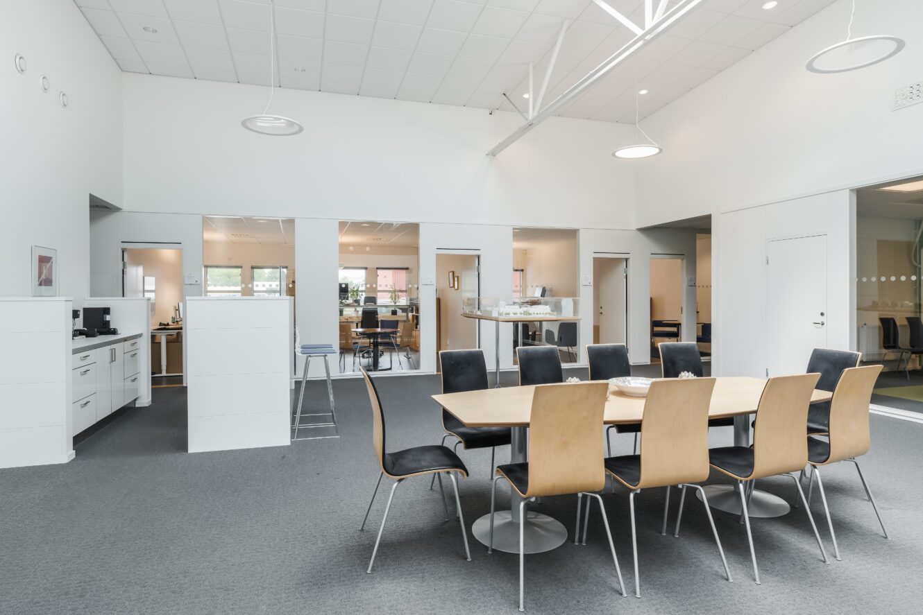 Modernt kontorslandskap med ett stort konferensbord i trä, svarta stolar i trä och flera rum med glasdörrar i bakgrunden med stark belysning.