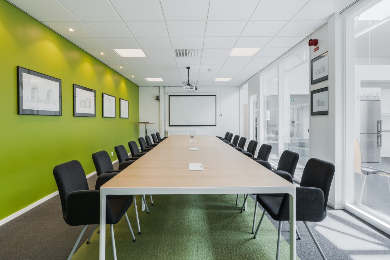 Modernt konferensrum med ett långt bord, svarta stolar, grön accentvägg, inramade tavlor, projektor och stora fönster som släpper in naturligt ljus.