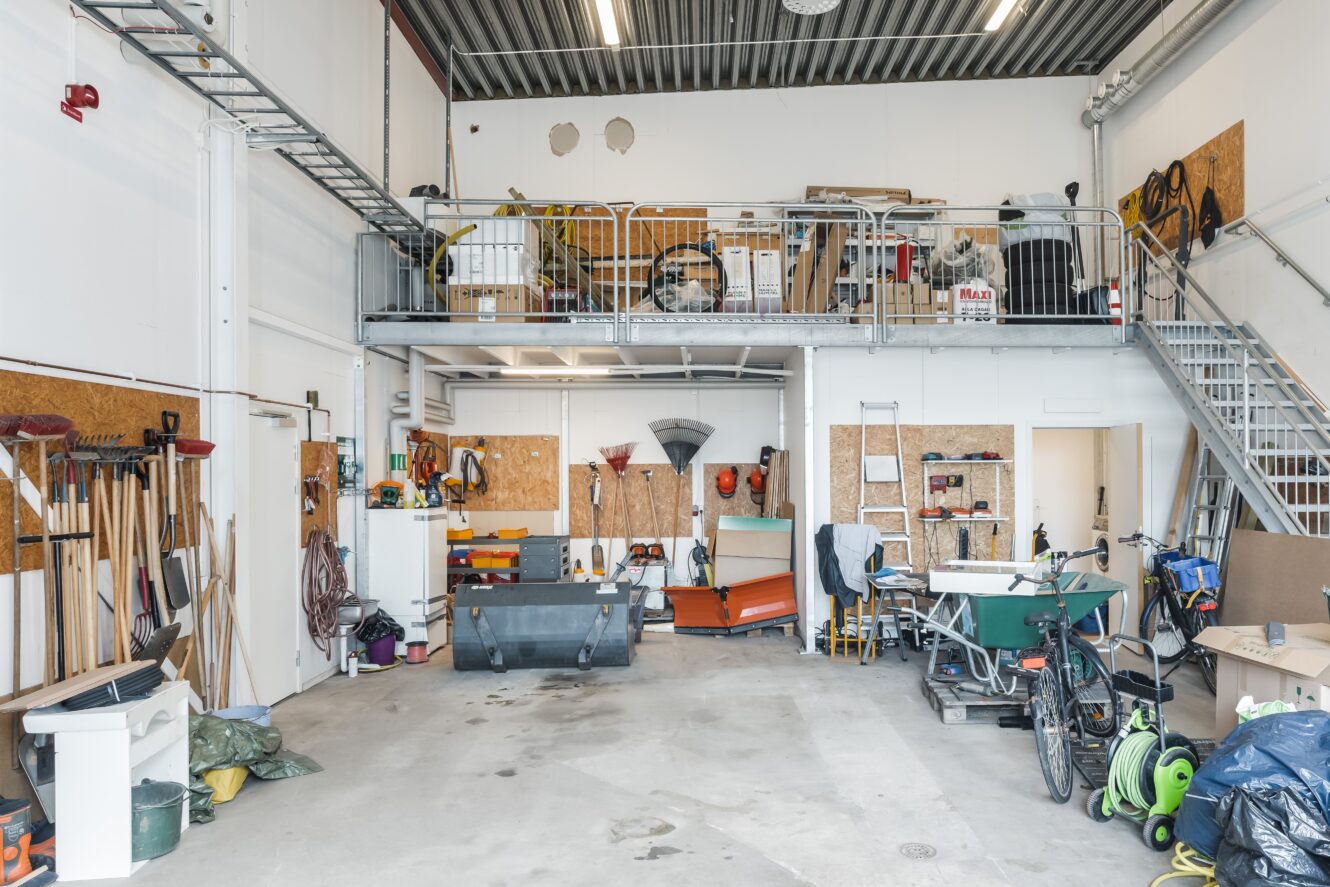 En rymlig verkstad med verktyg och utrustning organiserade längs väggarna, ett förvaringsutrymme på mezzaninen och en metalltrappa som leder till den övre nivån.