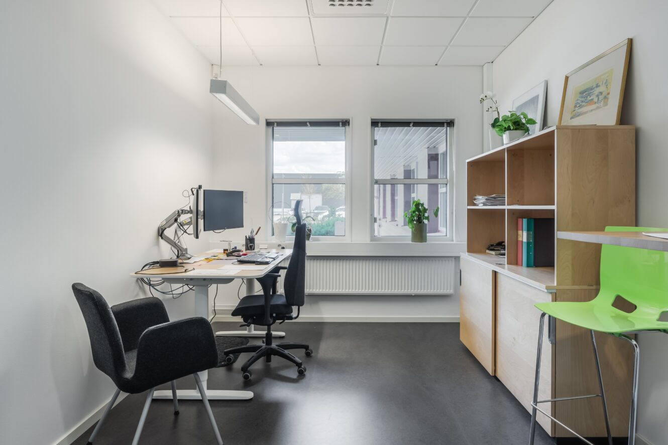 Ett modernt kontor med skrivbord, dator, svart stol, två fönster, hyllor med böcker och växter samt en grön stol.