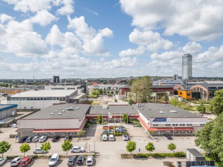 Flygfoto över ett affärsdistrikt med låga kontorsbyggnader, parkerade bilar, trädkantade gator och ett högt torn i bakgrunden under en delvis molnig himmel.