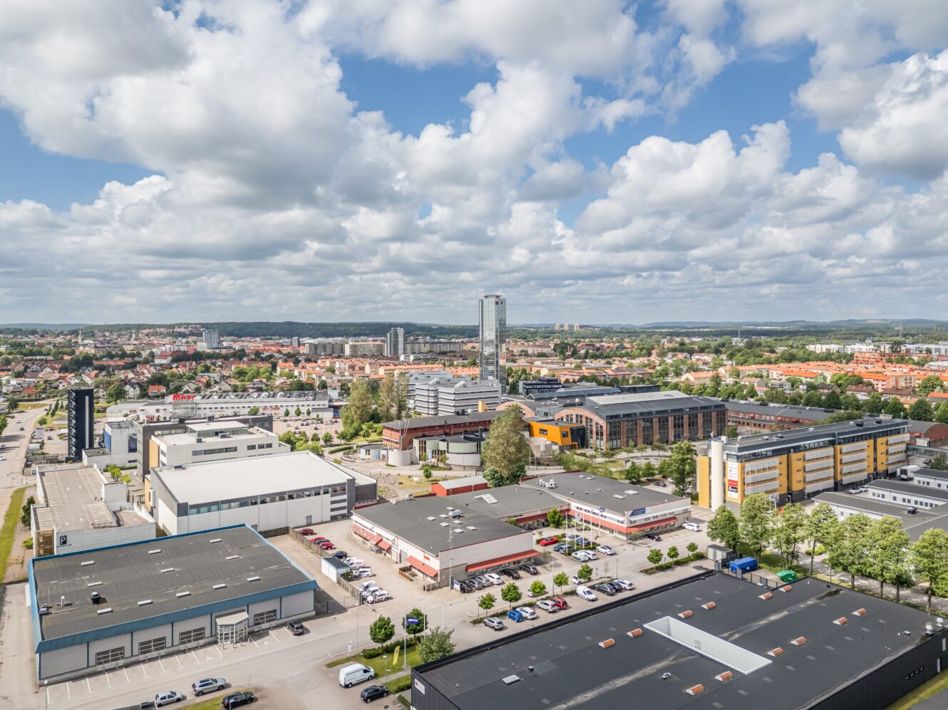 Flygfoto över ett industri- och handelsområde med lagerlokaler, kontorsbyggnader och en stadsbild i bakgrunden under en delvis molnig himmel.