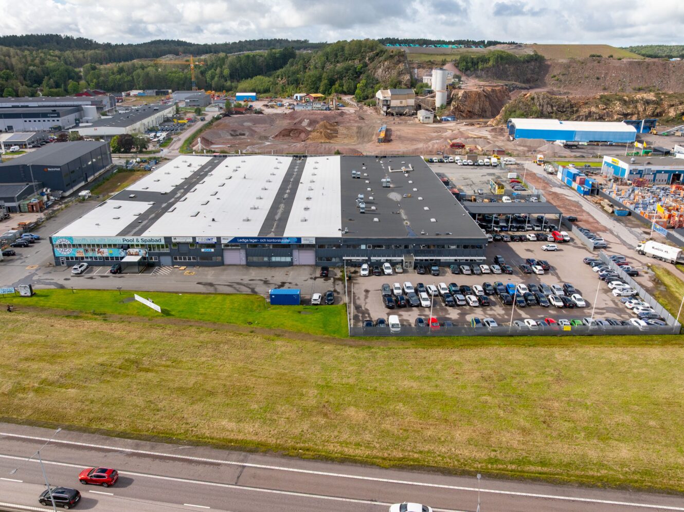 En stor industribyggnad med en parkeringsplats full av bilar, omgiven av vägar och byggarbetsplatser, med träd och kullar i bakgrunden.