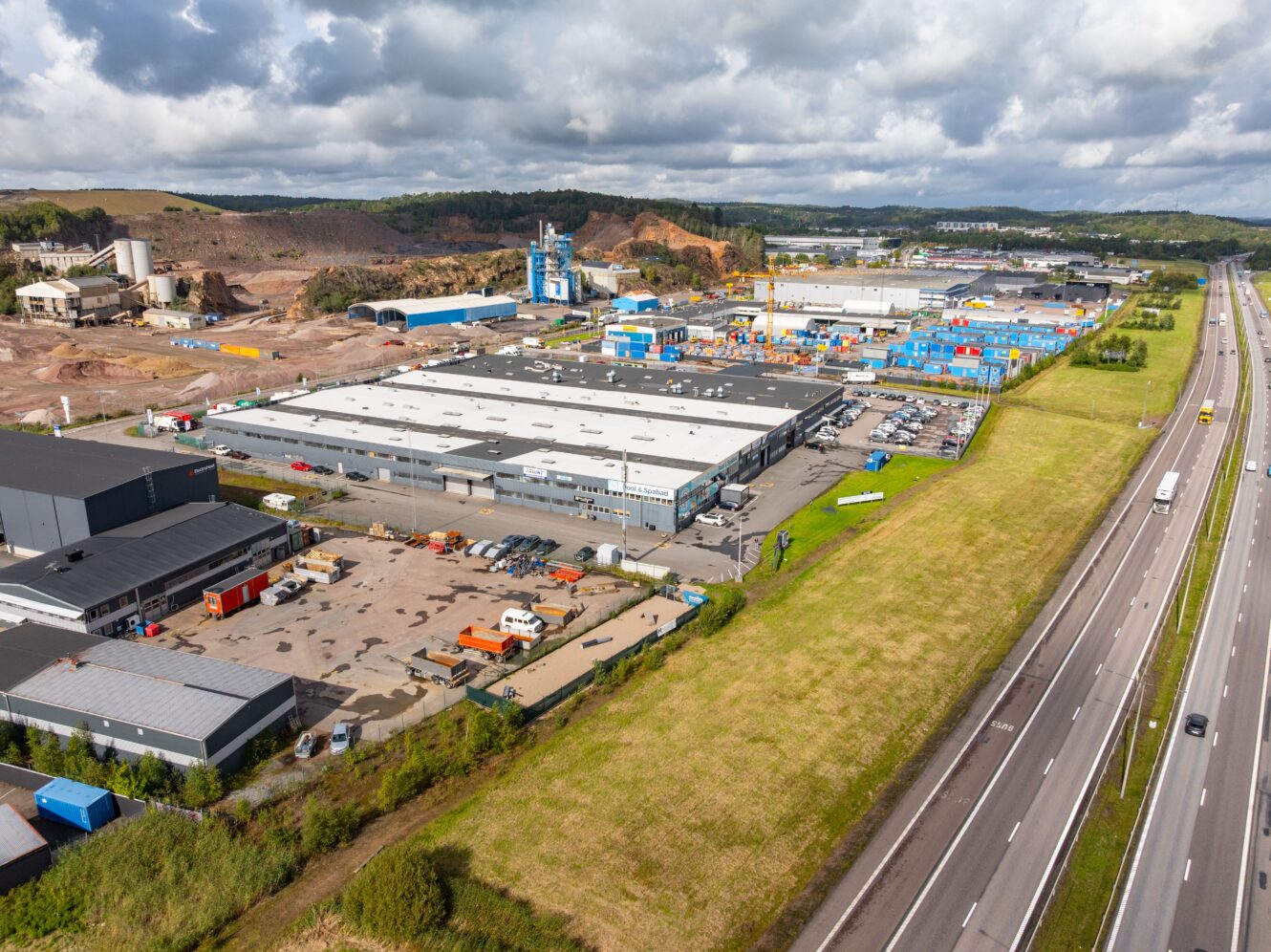 Flygfoto över ett industriområde med lagerlokaler och fabriker intill en motorväg, omgivet av gräs och kullar under en delvis molnig himmel.