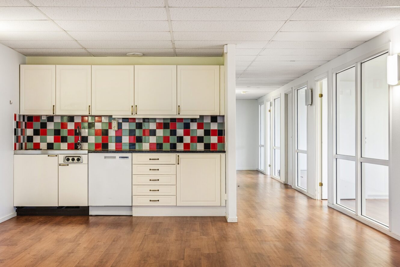 Ett litet kök med vita skåp, ett bakstycke med färgglada kvadratiska plattor, trägolv och stora fönster längs en hall.