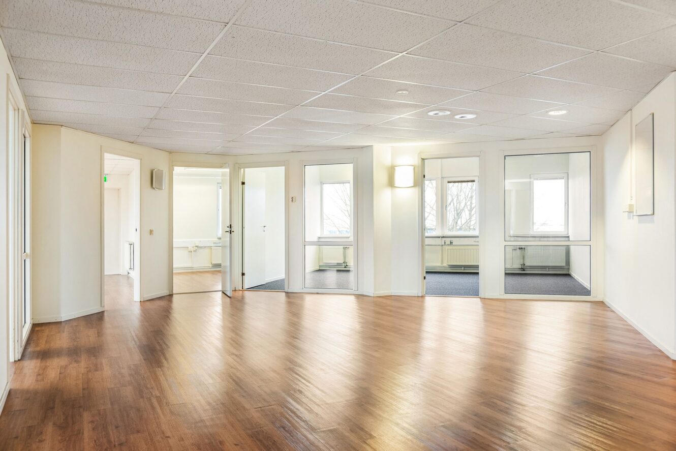 Rymligt tomt kontorsrum med ljusa väggar, trägolv, takplattor, glasdörrar och fönster som släpper in naturligt ljus.