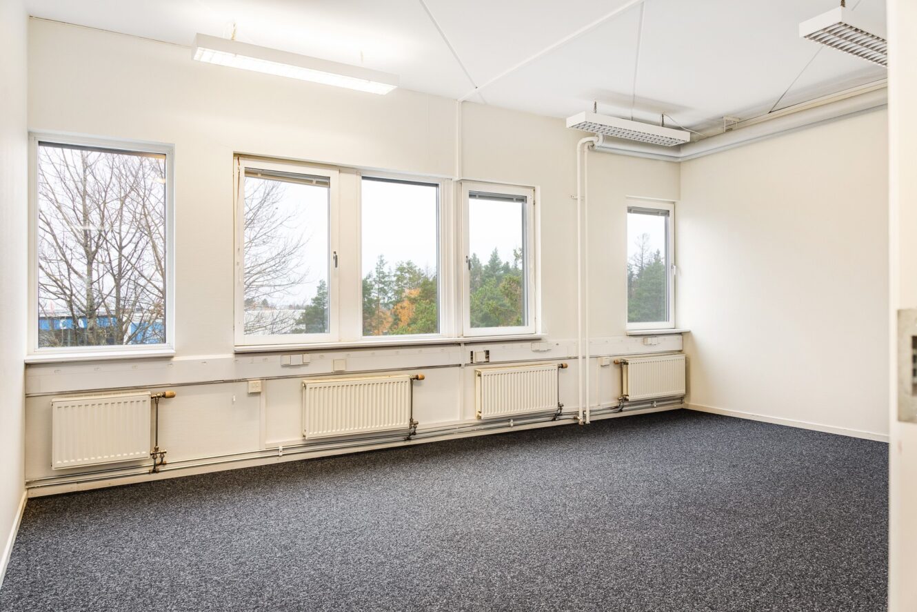 Tomt kontorsrum med tre stora fönster, vita väggar, grå heltäckningsmatta, lysrör i taket och synliga radiatorer under fönstren.