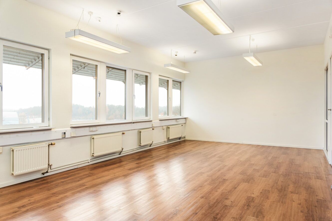 Ett tomt rum med vita väggar, stora fönster, trägolv och taklampor. Naturligt ljus kommer in genom fönstren längs ena sidan av rummet.