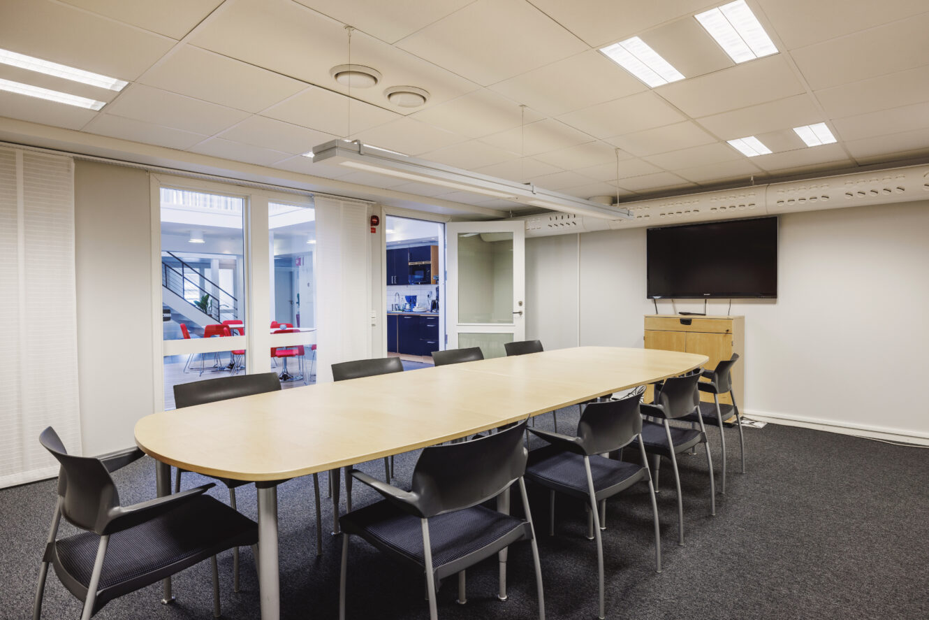 Ett modernt konferensrum med ett ovalt bord, tio svarta stolar, en väggmonterad TV och glasdörrar som leder till ett annat kontorsområde.